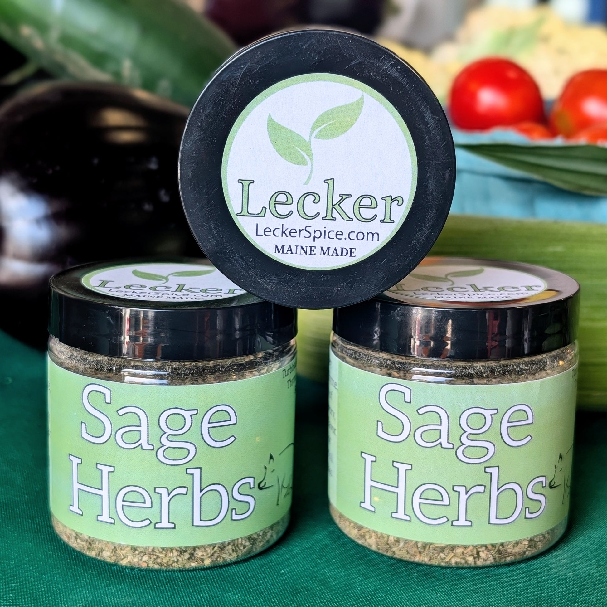 Two jars of Lecker Sage Herbs with black lids and light green labels, with one propped on top on its side with the lid showing.. The jars are set against a blurred background of fresh vegetables including tomatoes, eggplant, and cucumber 