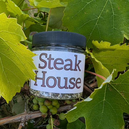 A small clear plastic jar with a black lid and a white label reading 'Steak House' in brown lettering, placed among green grape leaves and unripe grapes outdoors.
